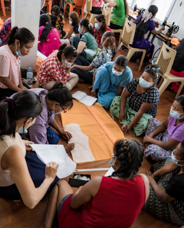 A group of women wearing face masks sit on the floor around a table, working on sewing or cutting fabric in a workshop.