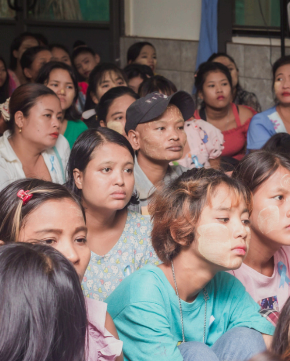 A crowd of people seated indoors, with several faces visible in the foreground and background. The scene appears to be a real environment with a wall and windows, so this is classified as Lifestyle rather than Product/Packaging.