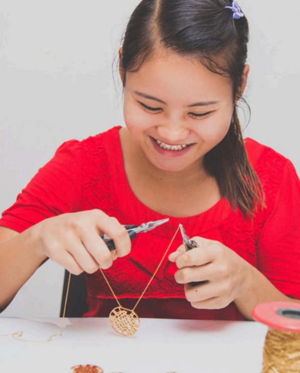 A smiling woman in a red top uses pliers to manipulate a small gold circular wire craft piece on a white surface.