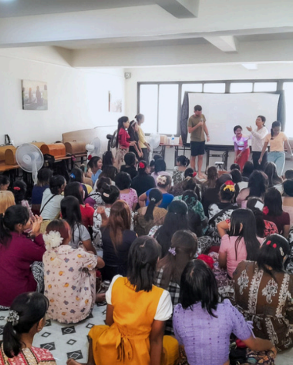 A large group of people, mostly women, are seated on the floor in a classroom or community hall, facing a presenter standing near a whiteboard and windows.