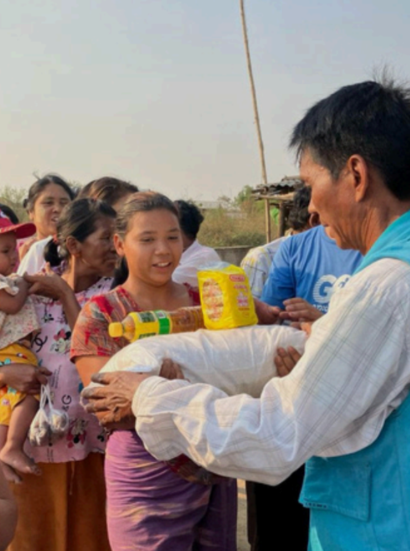 A man in a light blue vest and white shirt receives a white sack of rice from a woman in a patterned top and purple skirt, with several people and yellow bottles visible in an outdoor setting.