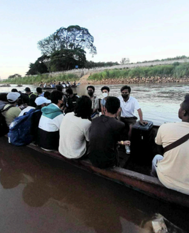 A group of people sitting in a narrow wooden boat on a river, with a grassy bank and trees in the background.