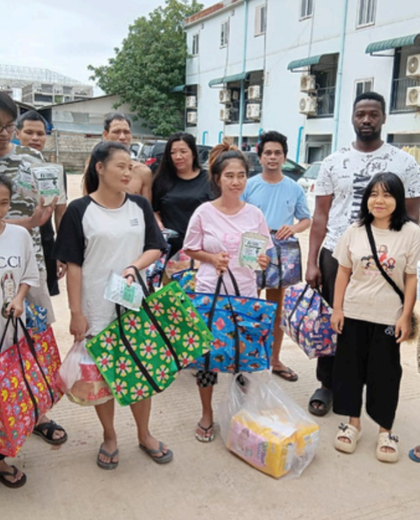 A group of nine people standing outdoors on a paved area, each holding or carrying a colorful reusable tote bag. The bags feature various floral and geometric patterns in green, blue, pink, and purple, with black handles.