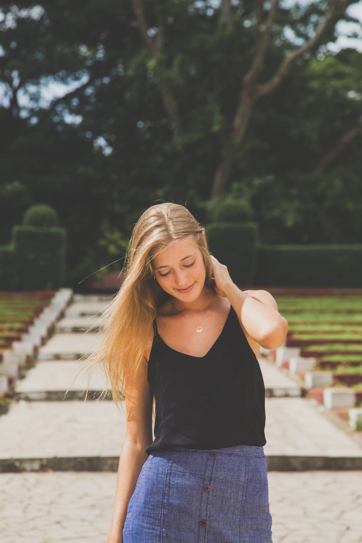 Woman with long blonde hair standing outdoors on a staircase with greenery in the background