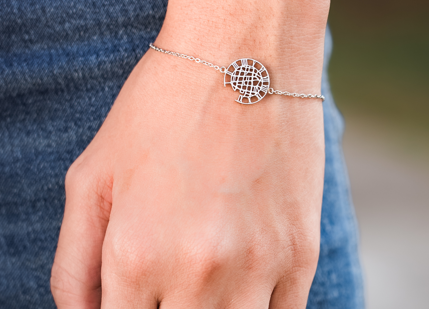A close-up of a person's hand wearing a delicate silver chain bracelet with a circular city map design charm.