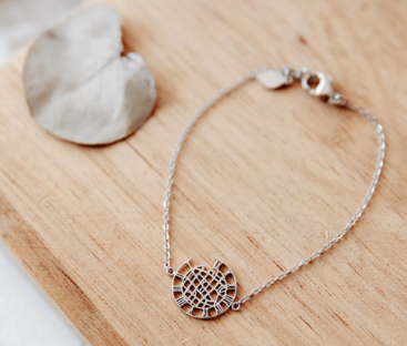 A delicate silver chain bracelet with a geometric, web-like circular pendant, displayed on a light wood surface next to a dried flower petal.